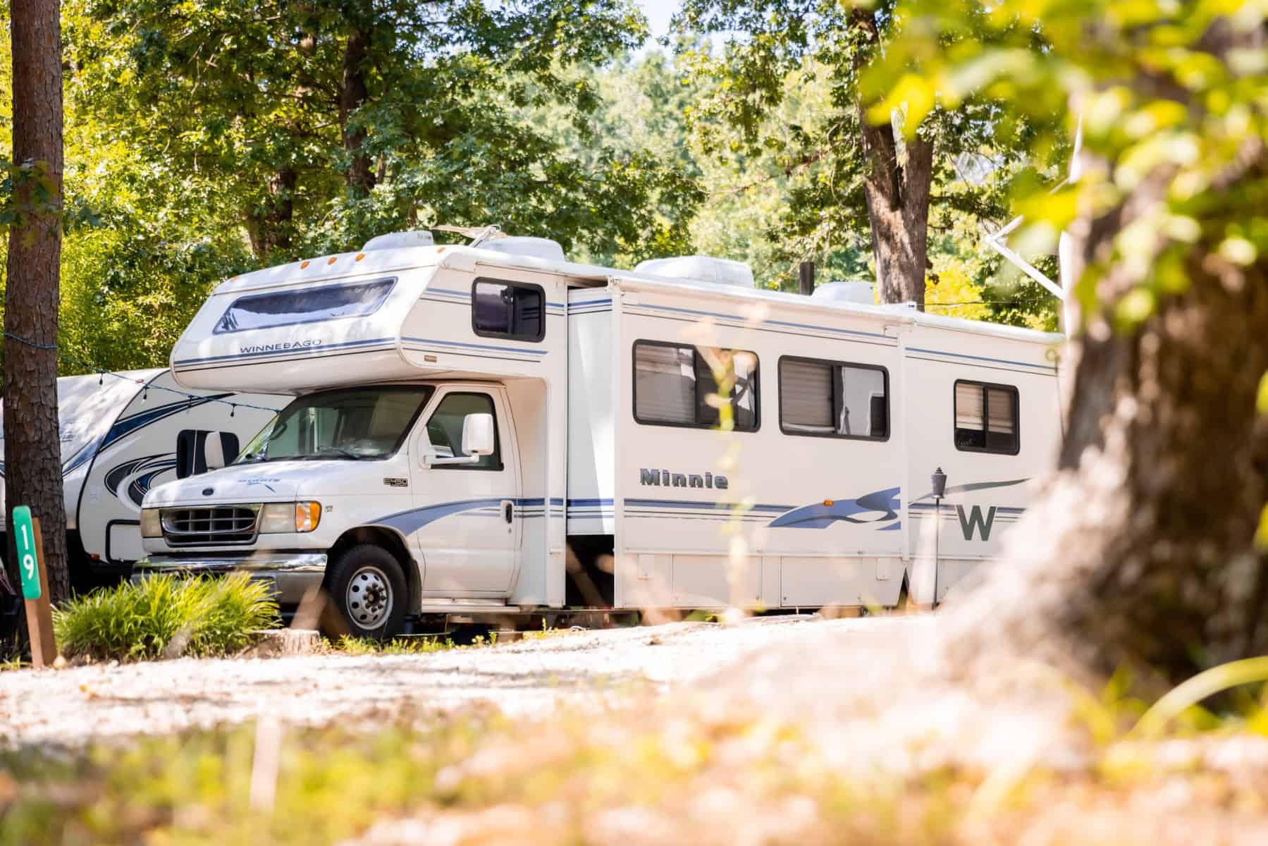 A white motorhome nestled in a wooded campsite feels like a home away from home. Sunlight filters through the trees, casting dappled shadows on the RV. The lush grass and foliage create a serene outdoor setting, making it an idyllic escape into nature.