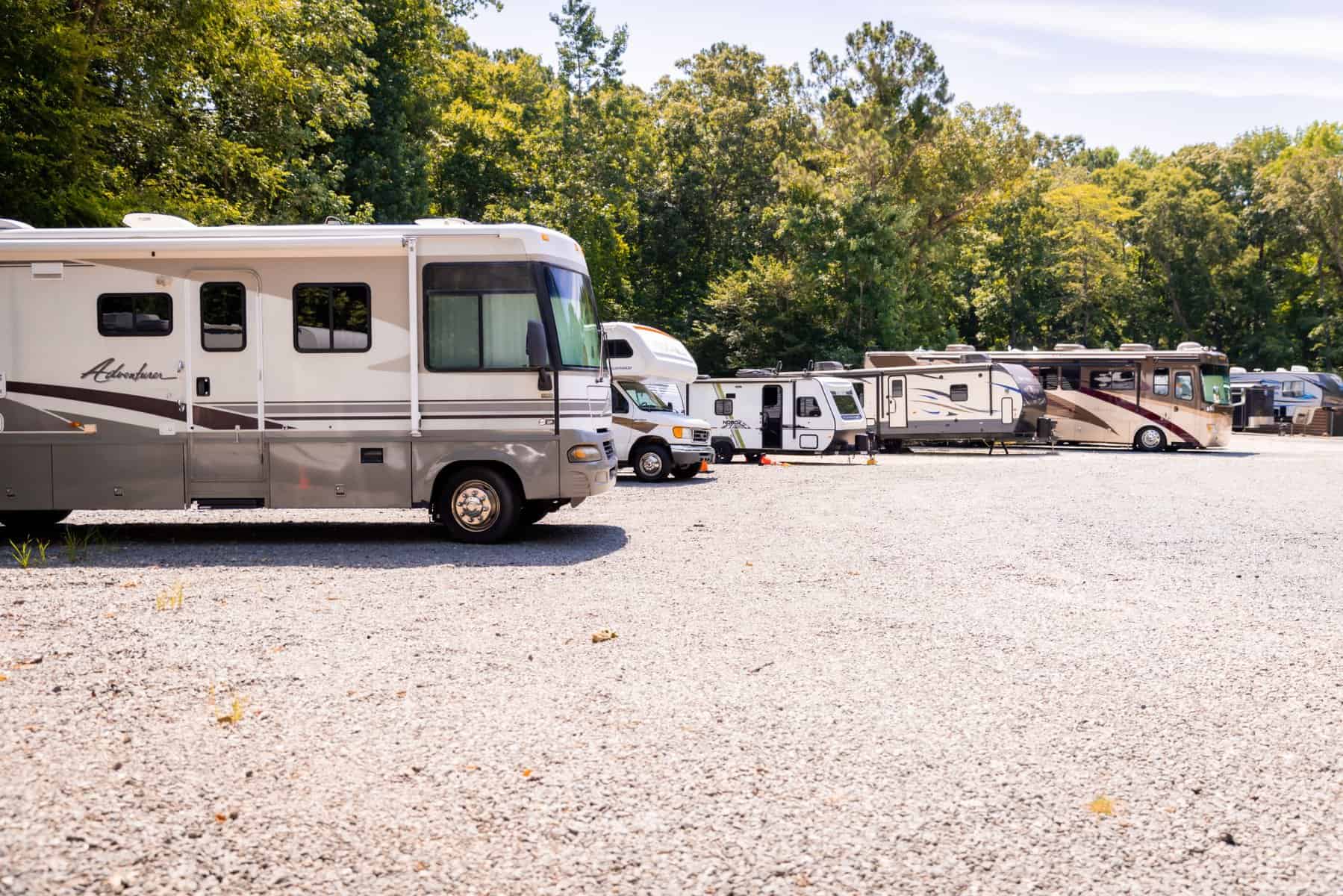 A row of parked RVs on a gravel lot surrounded by trees feels like a home under a clear blue sky.