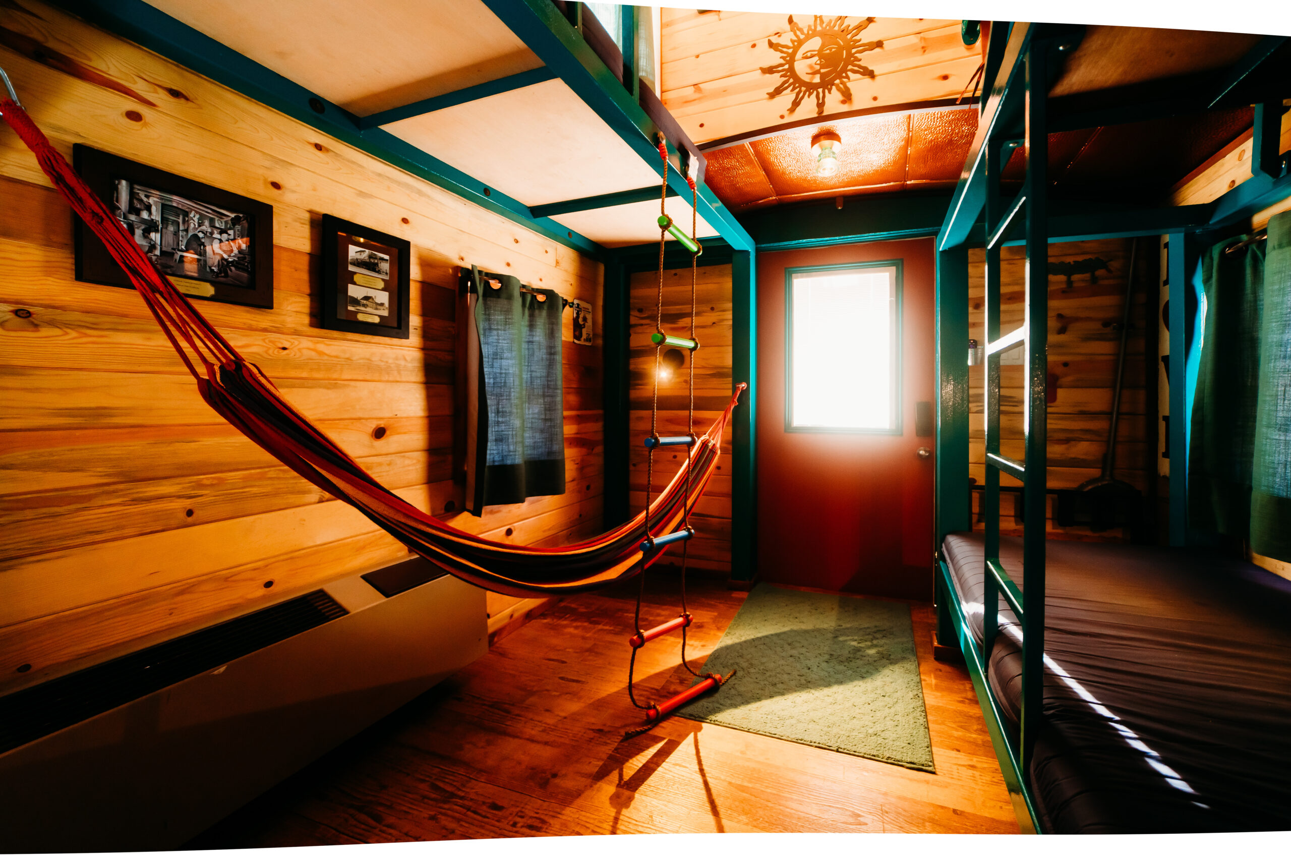 A cozy wooden room with a hammock, bunk beds, framed pictures on the wall, a sun decoration on the ceiling, and sunlight streaming through a door with a window.