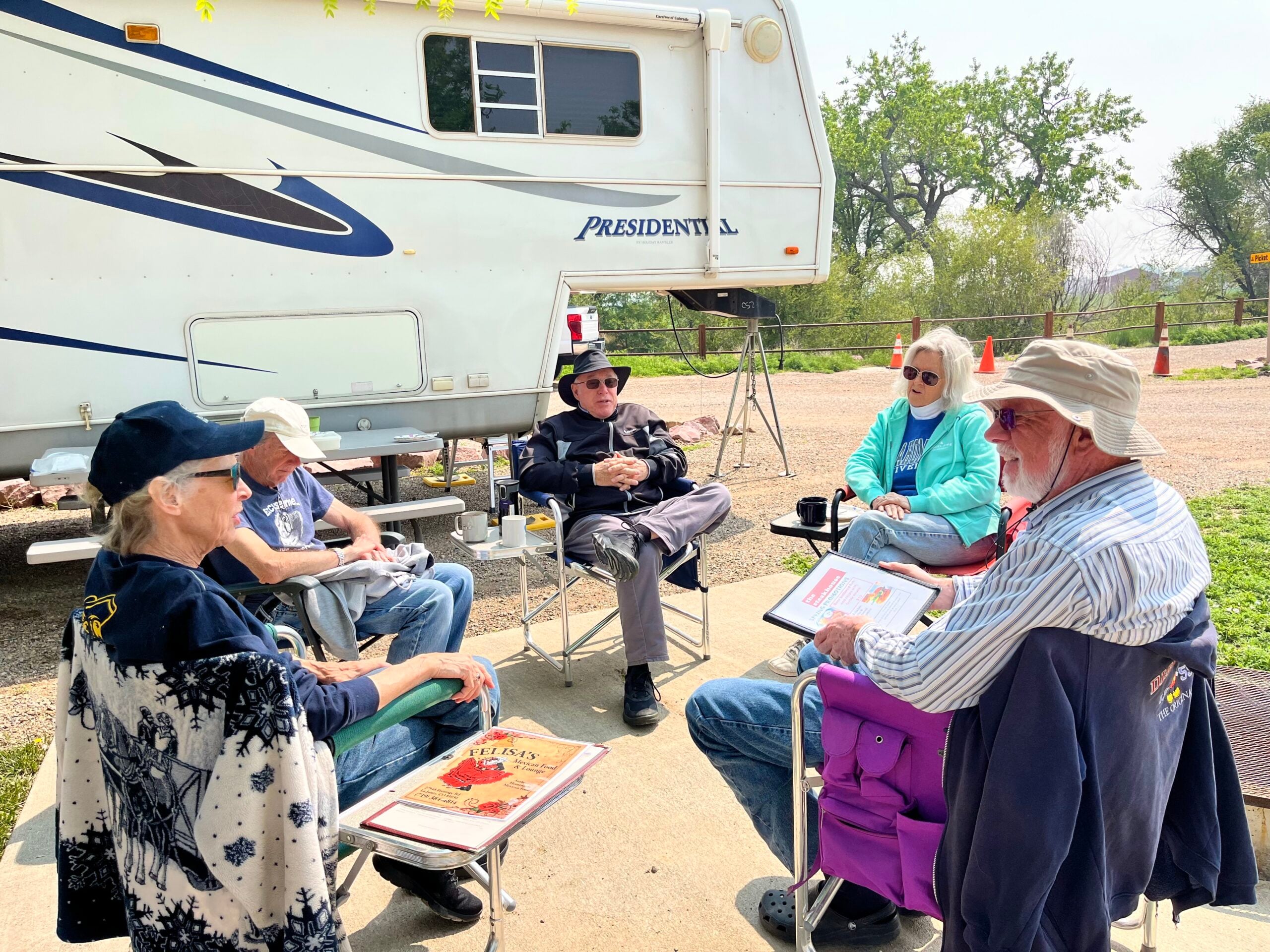 Five older adults sit in a circle on folding chairs outside an RV, talking and holding papers or books on a sunny day.