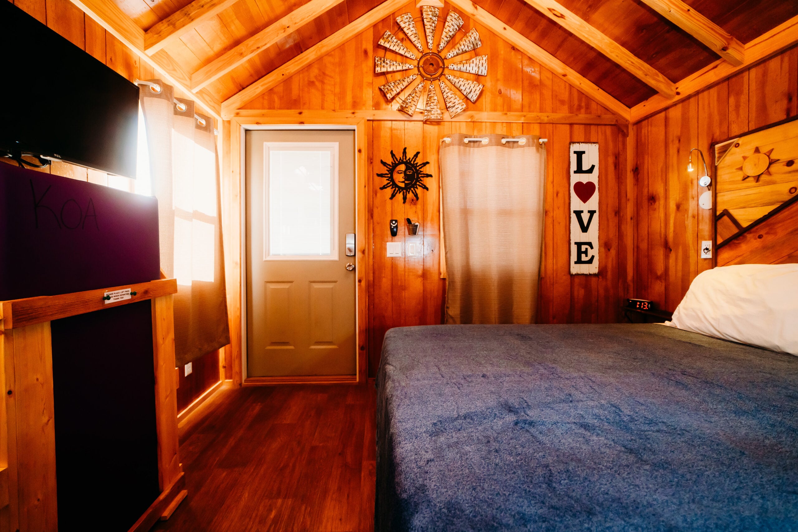 A small wooden cabin room with a bed, TV, door, and wall decorations including a "LOVE" sign and sun-shaped ornament; sunlight streams through window blinds.