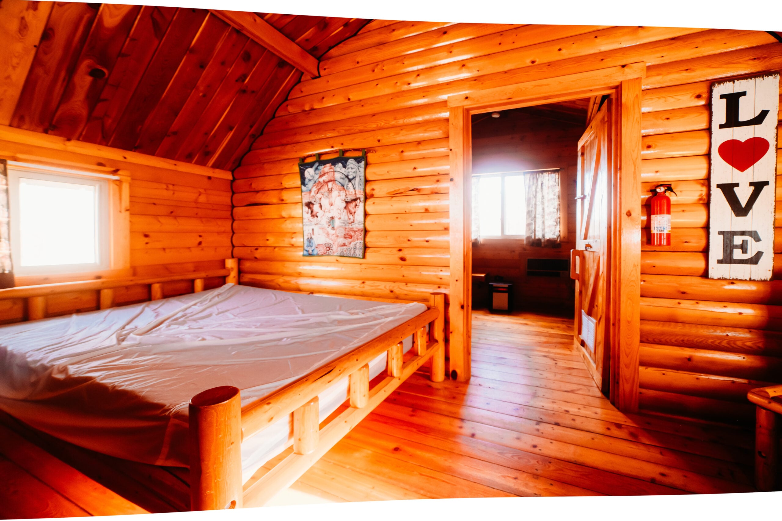 Interior of a wooden log cabin bedroom with an unmade bed, a LOVE sign on the wall, and sunlight streaming through windows in the adjacent room.