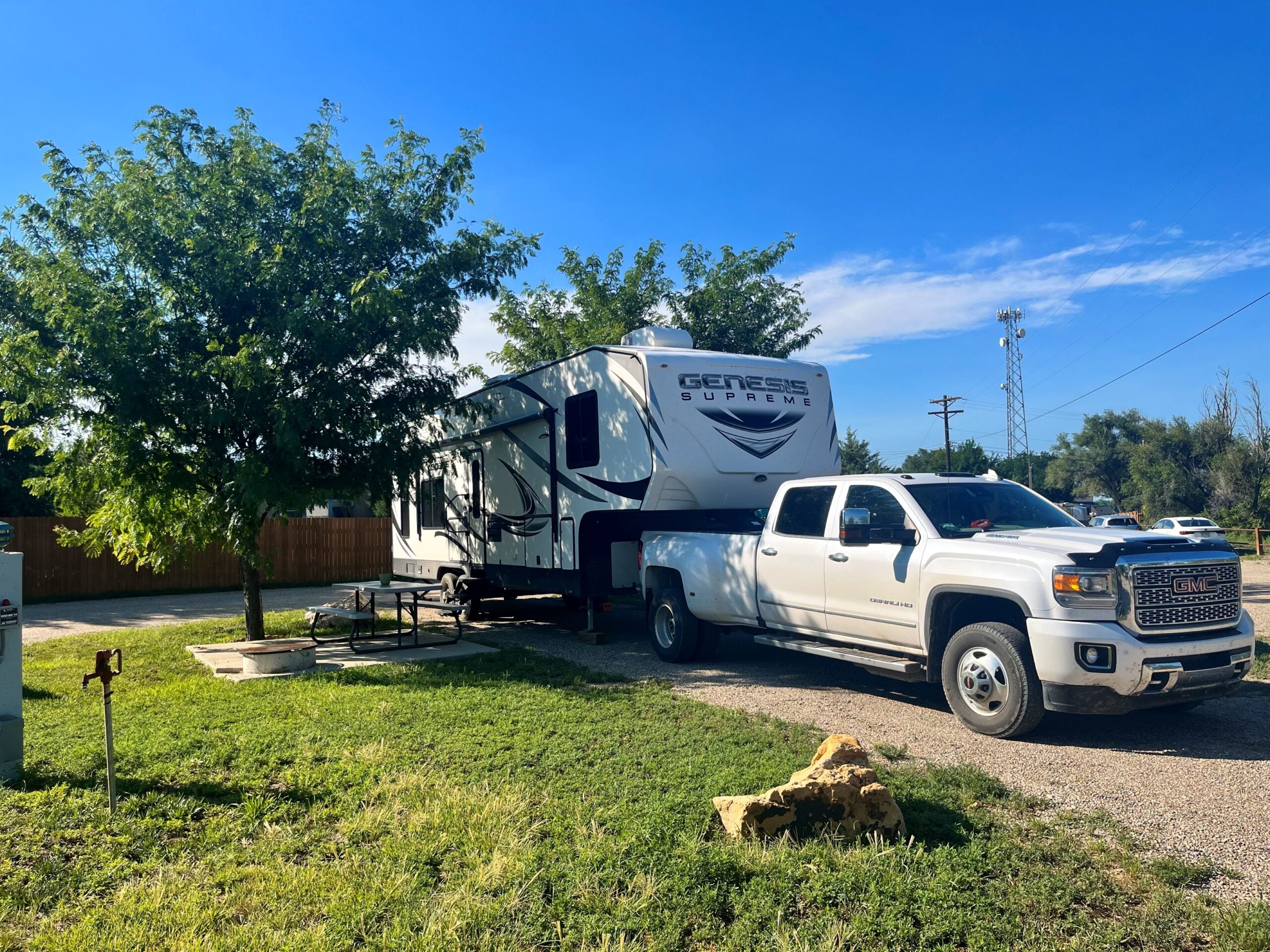 A white pickup truck is parked next to a Genesis Supreme travel trailer at a campsite with a picnic table, tree, and grassy area under a clear blue sky.