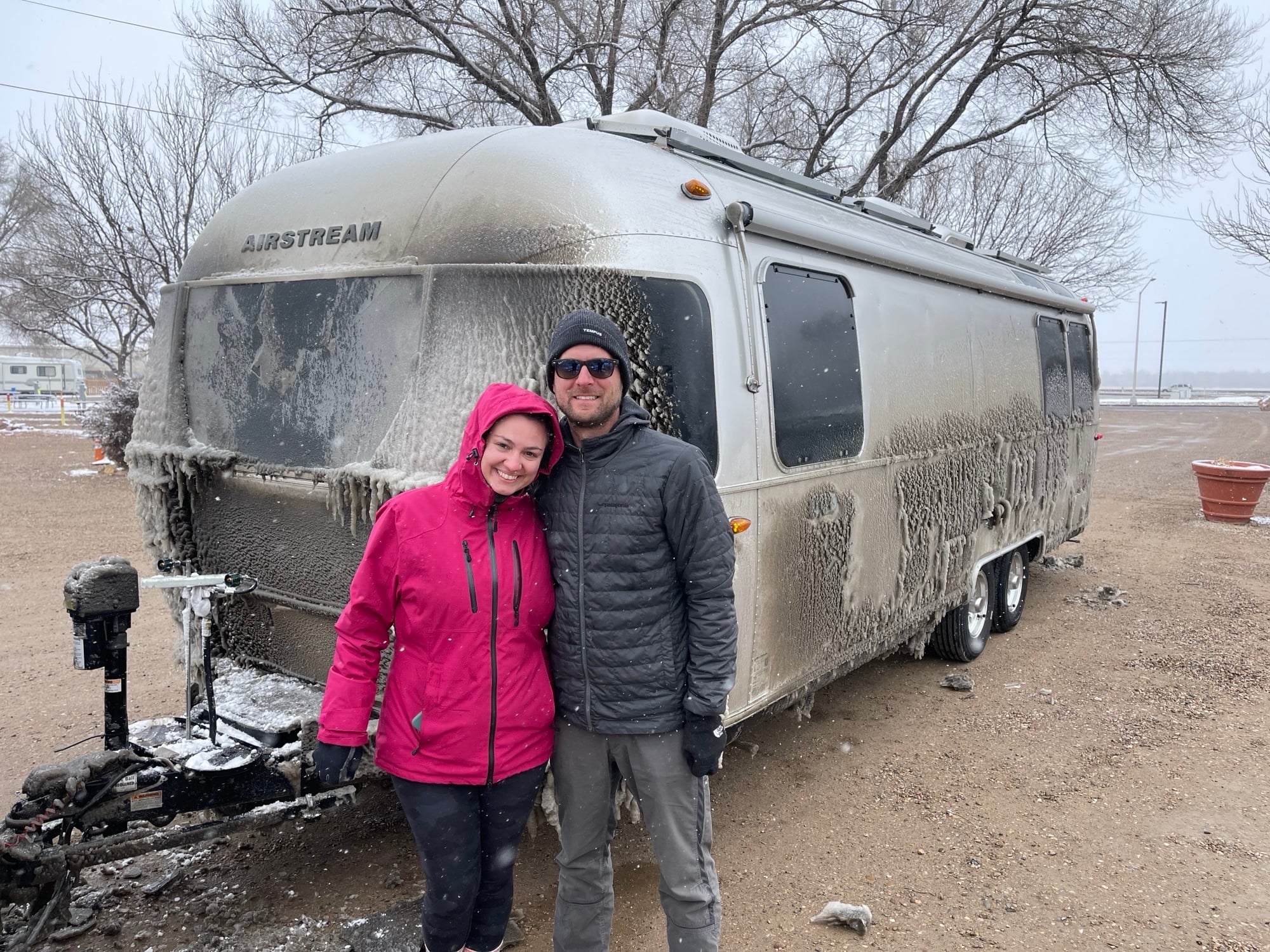 Two people in winter jackets stand in front of an Airstream trailer covered in ice and snow, with bare trees and overcast skies in the background.