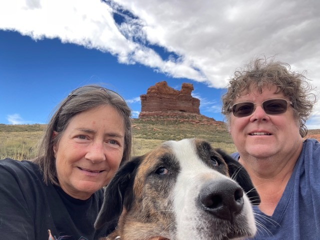 A woman and a man take a selfie with a dog in front of a red rock formation.